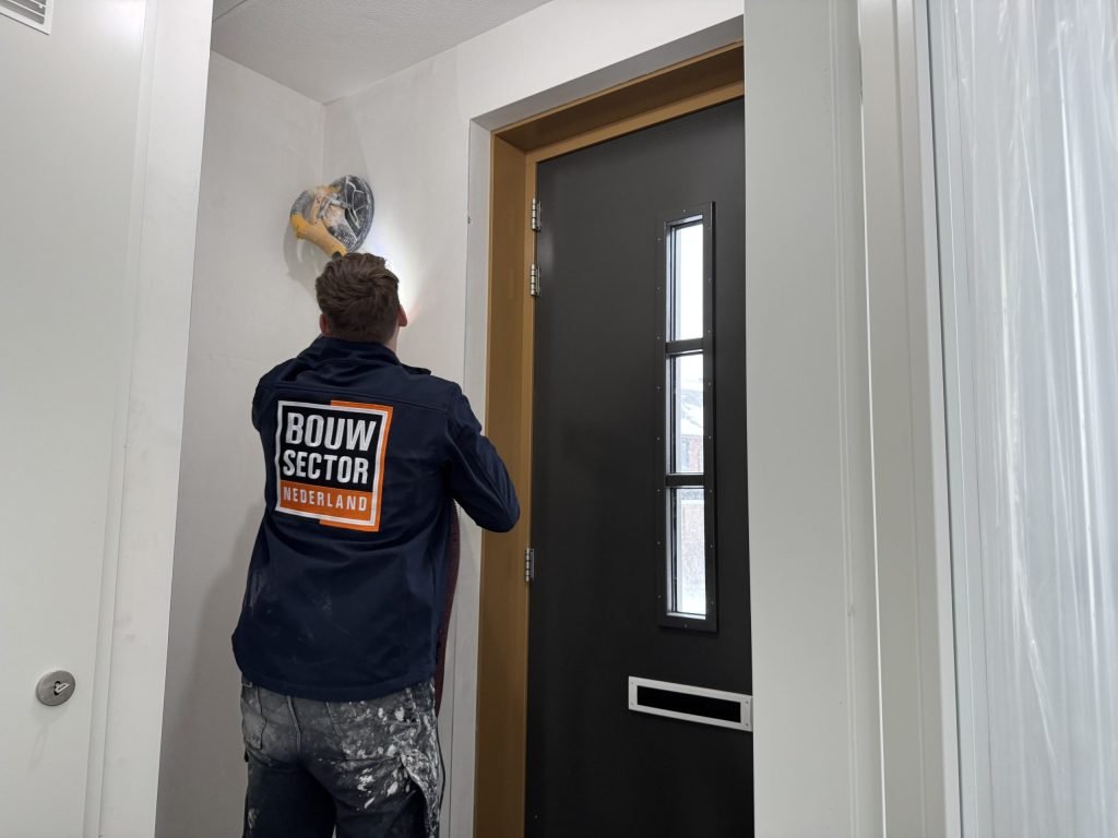 Vakman schuurt bij het plafond in de hoek van een hal, naast de voordeur