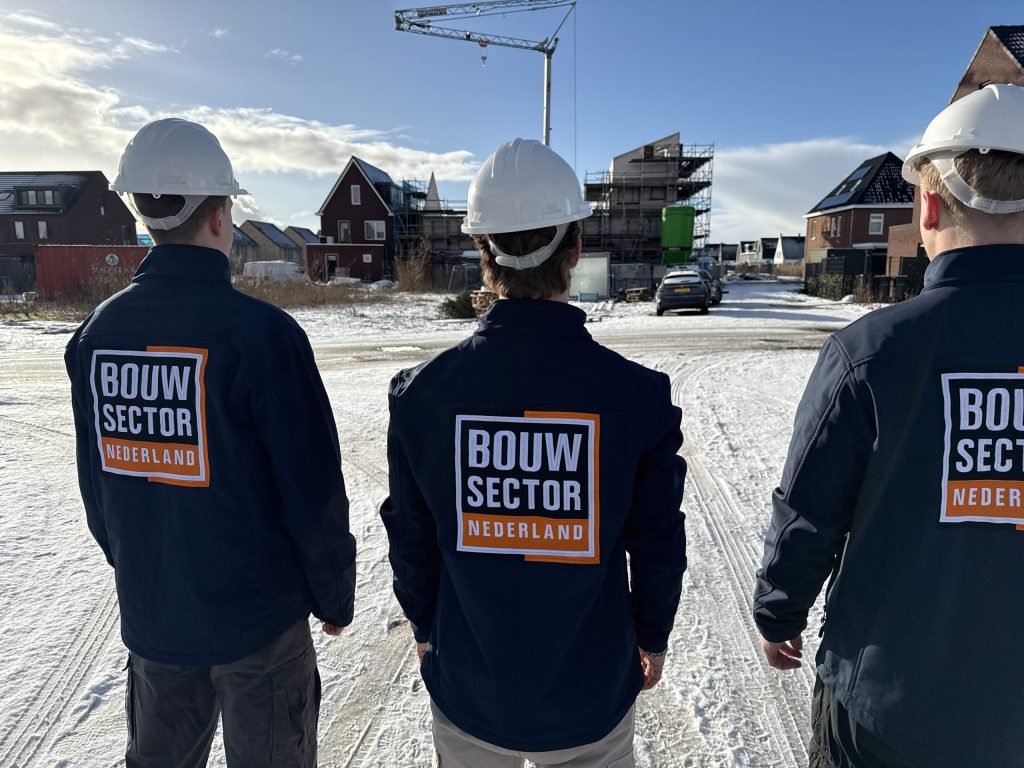 Drie vakmensen van Bouwsector Nederland met witte helm kijken uit over een straat met bakstenen woningen in aanbouw