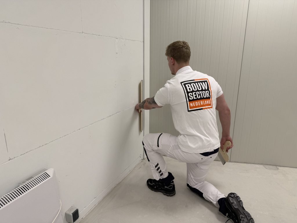 Stukadoor zit op één knie en werkt met een spaan de onderkant van een wand bij de hoek af